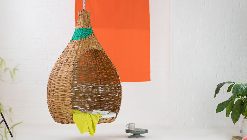 Hanging willow woven pod chair with a band of bright, colourful  recycled waste materials woven in to the design. Hanging in a bright, contemporary room with an orange background.  